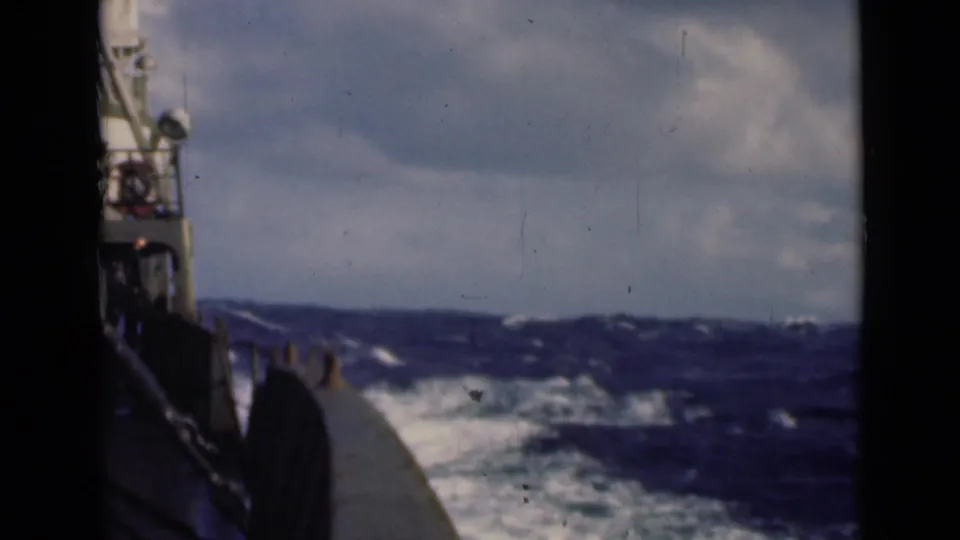 Deck-level view from a vessel cutting through rough seas, with railings, wake, whitecaps, and cloudy sky visible.