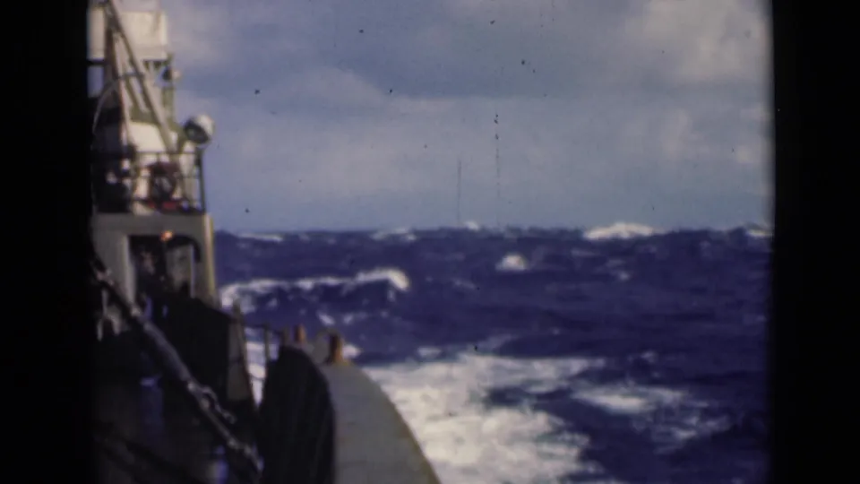 Aboard-ship view of turbulent open water, with vessel structure at left and foamy wake trailing alongside.