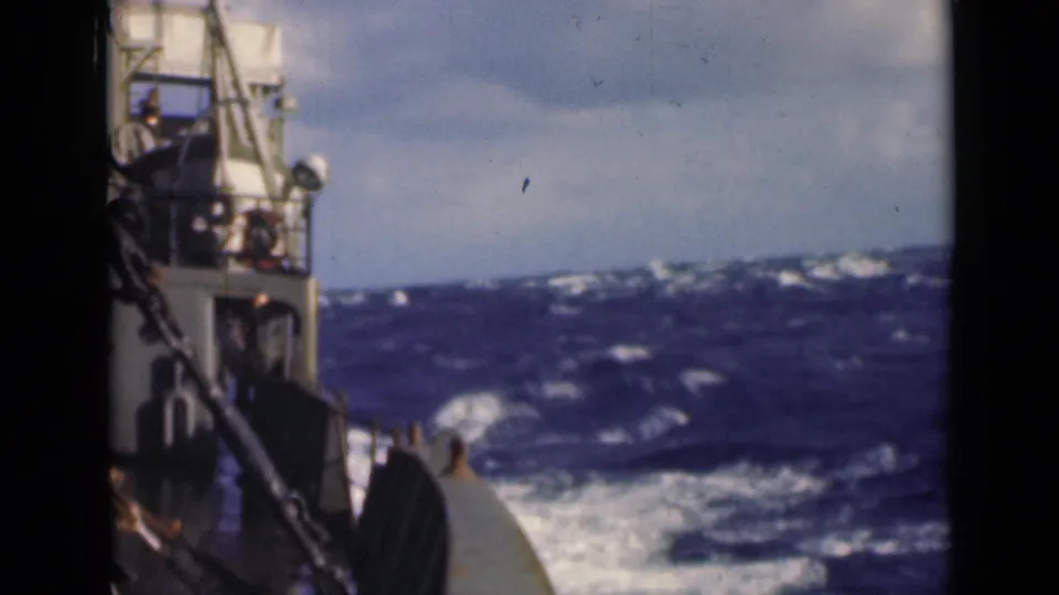 Handheld 8mm view from a vessel at sea, showing deck rail, mast structure, rough waves, and a cloudy horizon.