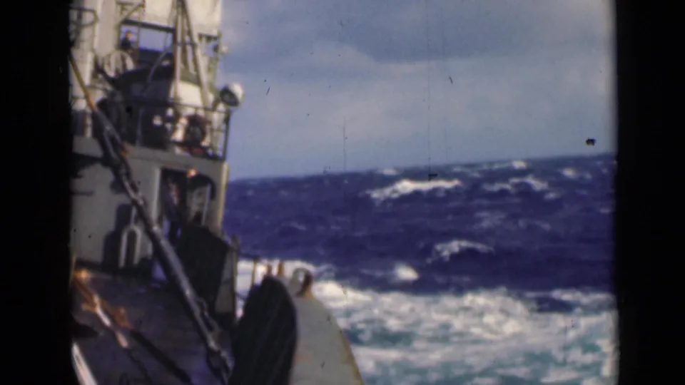 The vessel's deck edge and rail pass through turbulent water, with white wake and distant waves under cloudy light.