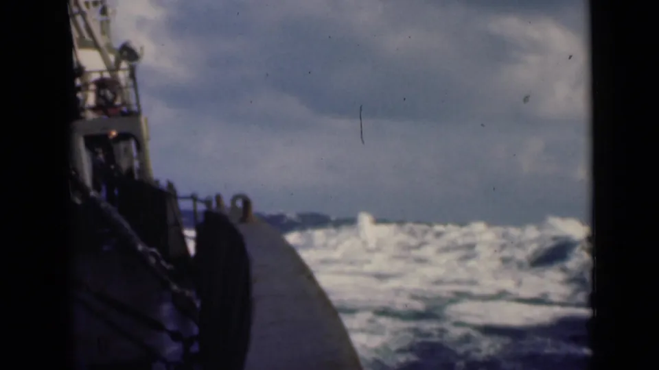8mm frame of a boat pushing through rough open water, with side railings, bow hardware, and overcast sky visible.