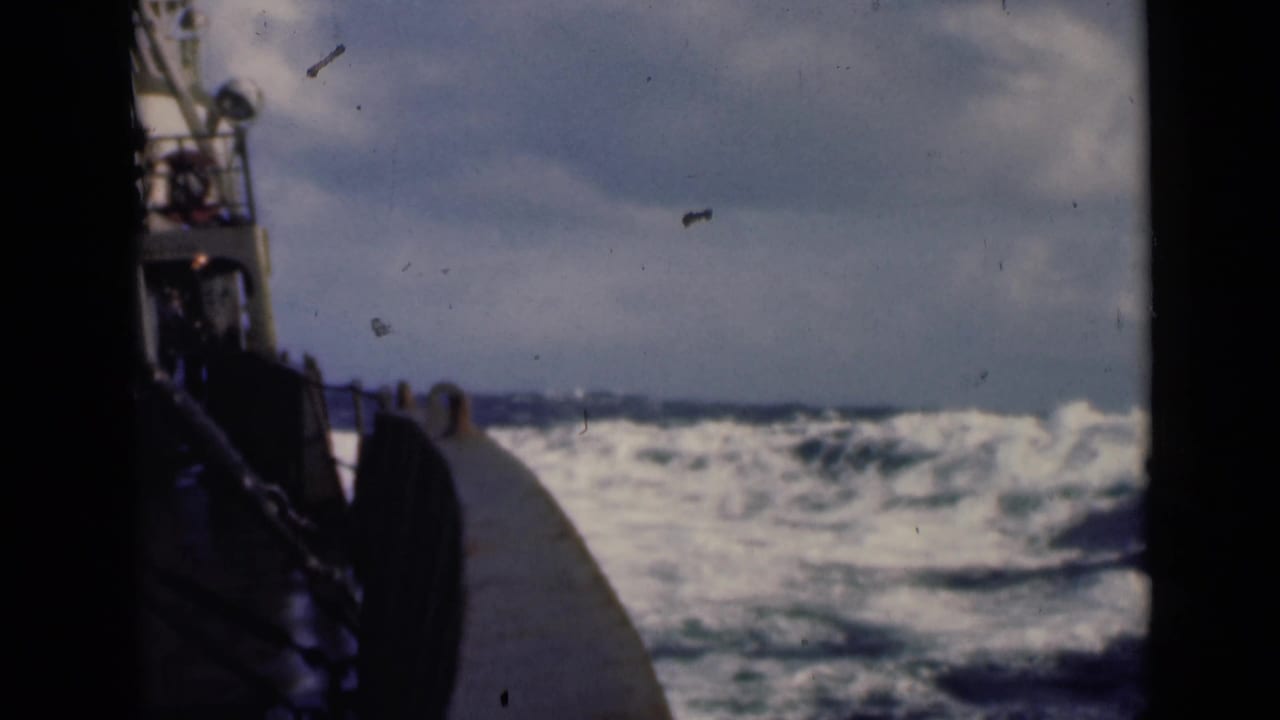 Ship deck edge beside rough ocean waves under a cloudy sky.