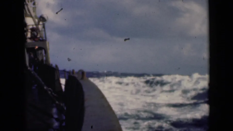 1940s 8mm frame from aboard a ship, looking past the deck edge toward churning waves and a cloudy horizon.