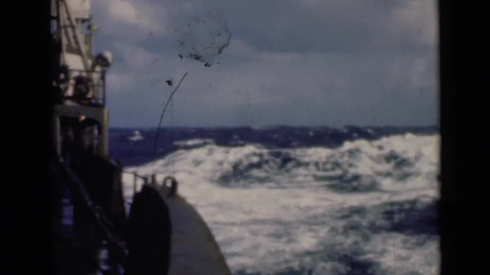 A shipboard 8mm view shows dark swells and white breaking waves beyond part of the vessel.