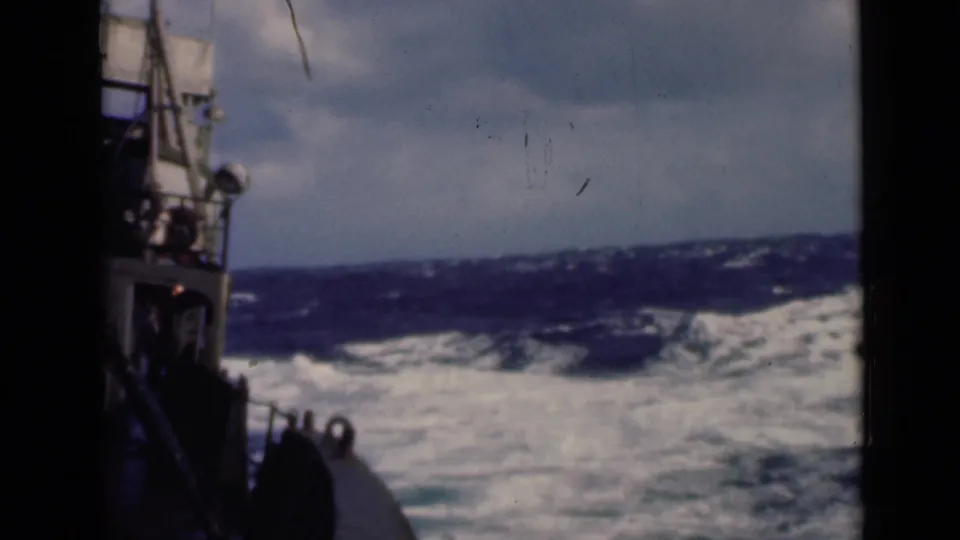 The vessel appears to pitch beside rough water, with white foam and dark blue swells visible through soft 8mm color.