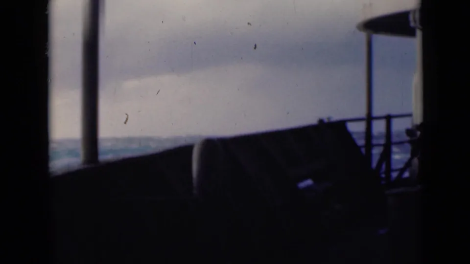 A darker shipboard frame shows railing or deck equipment silhouetted against distant open water and a cloudy sky.