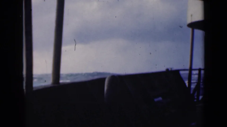 The camera looks past dark boat posts and a low rail toward an overcast horizon and churning water in archival 8mm color.