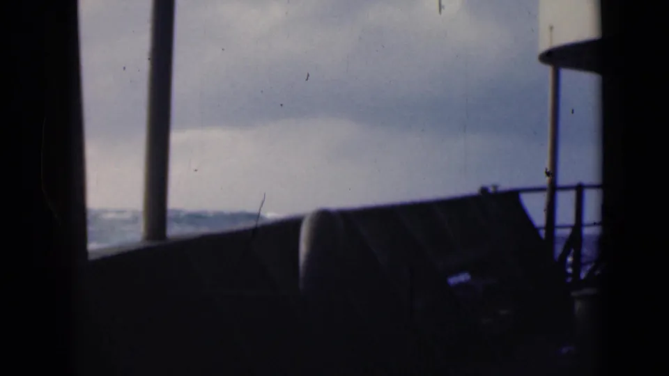 A dim 8mm frame shows rough water and a clouded horizon viewed through silhouetted posts and railings inside a boat.