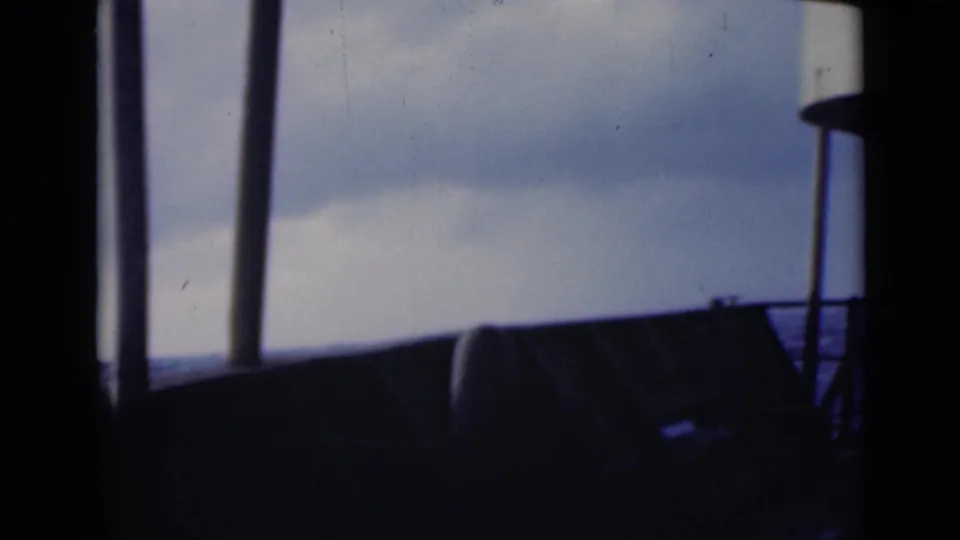 Handheld 8mm shipboard frame with dark window supports, a railing, and partial deck equipment under overcast sky.