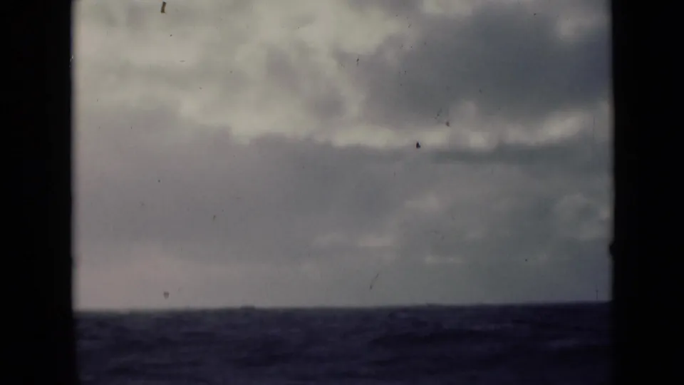 Archival 8mm frame showing a low ocean horizon beneath dense cloud cover, with visible dust and gate edges.