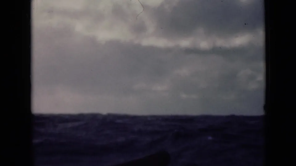 Rough open water under a clouded sky, framed by dark vertical ship-side shapes in faded 1947 8mm footage.