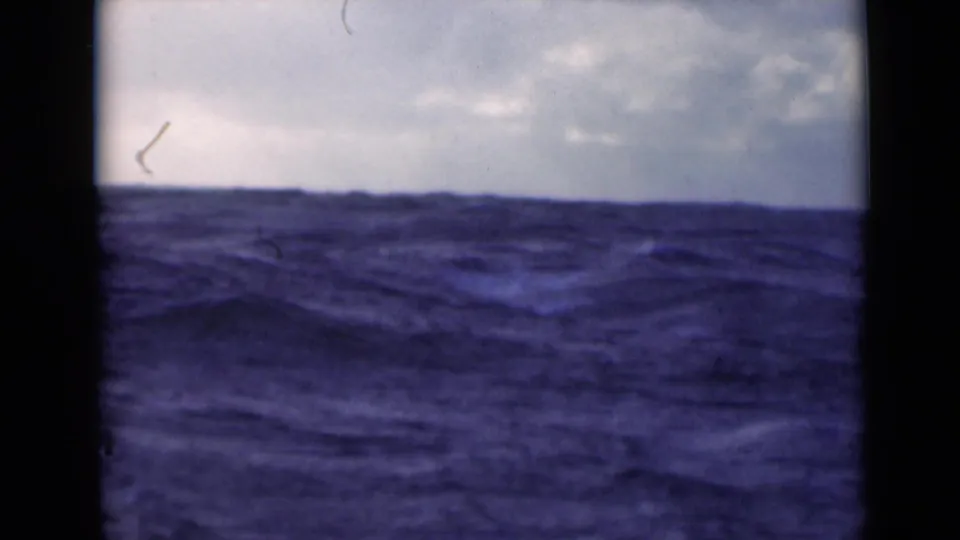A low horizon of rough blue water stretches under a pale cloudy sky, with dust and edge shadows from the 8mm film scan.