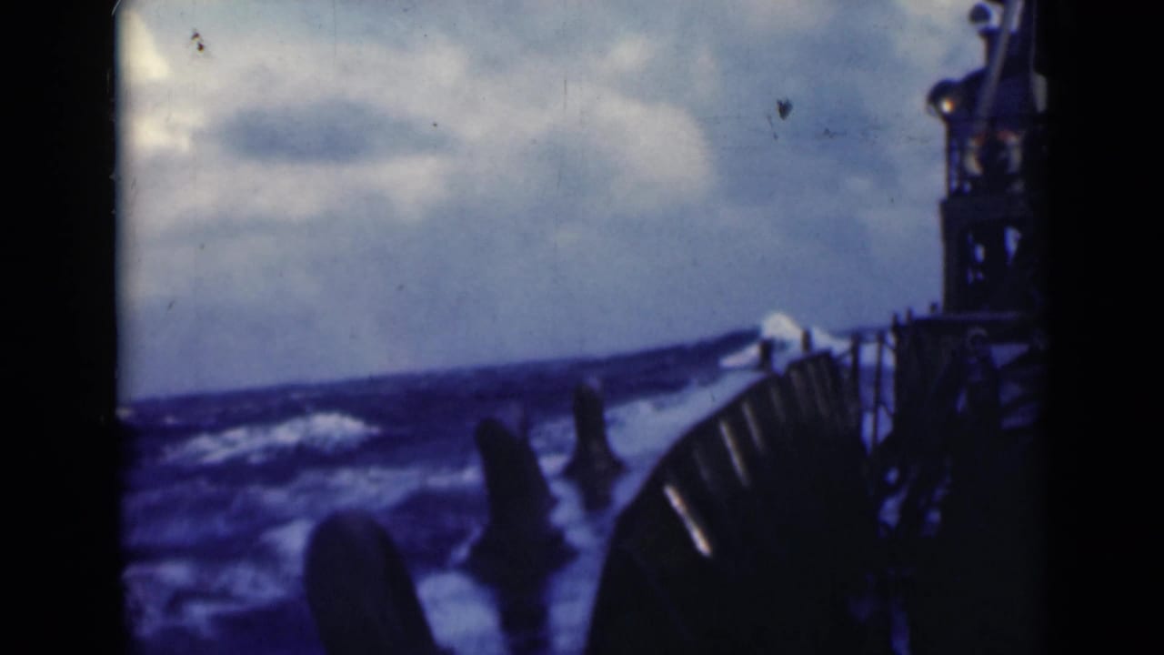 Turbulent ocean surface beside a ship railing with dark structure at right and cloudy sky above.