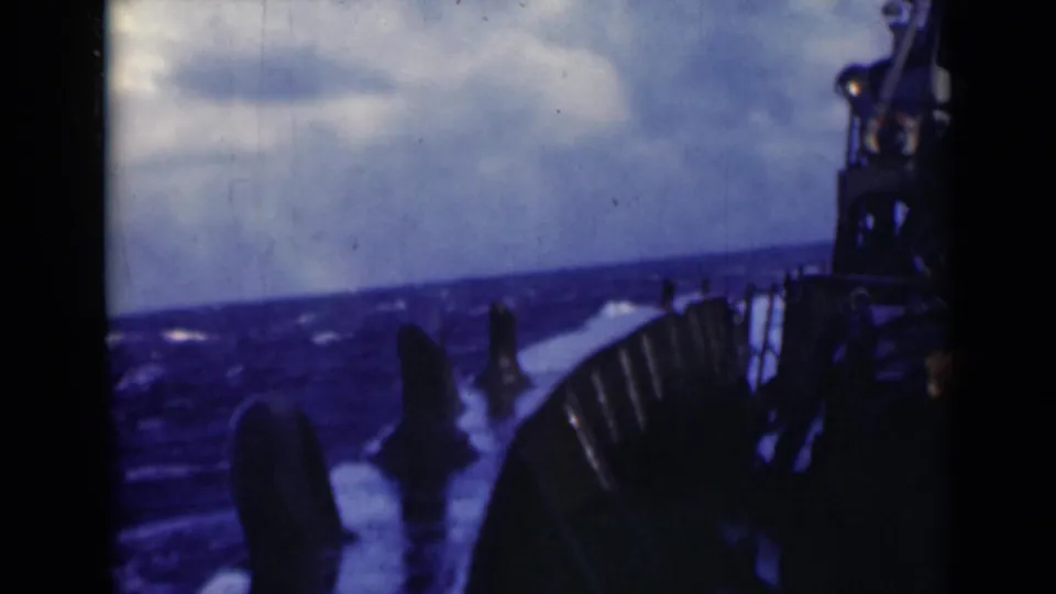 Handheld 8mm view from the vessel shows rough blue water, foam, railings, and silhouetted deck equipment in cloudy light.