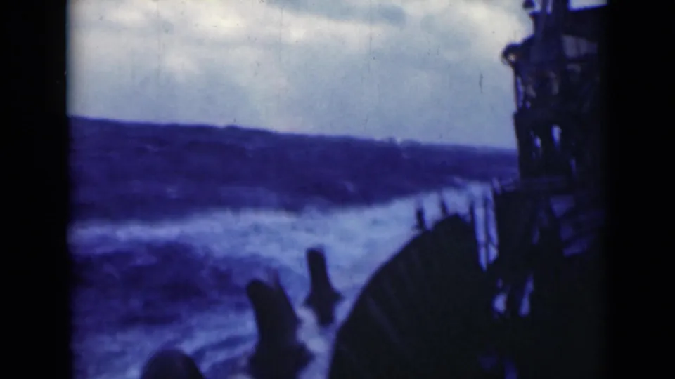 A larger swell passes alongside the ship, with foaming water, silhouetted railing, and dark vertical deck structure visible at frame edge.