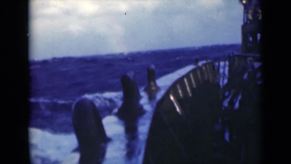 The vessel appears to pitch beside choppy blue water, with deck railings, ship fittings, and white spray filling the foreground.