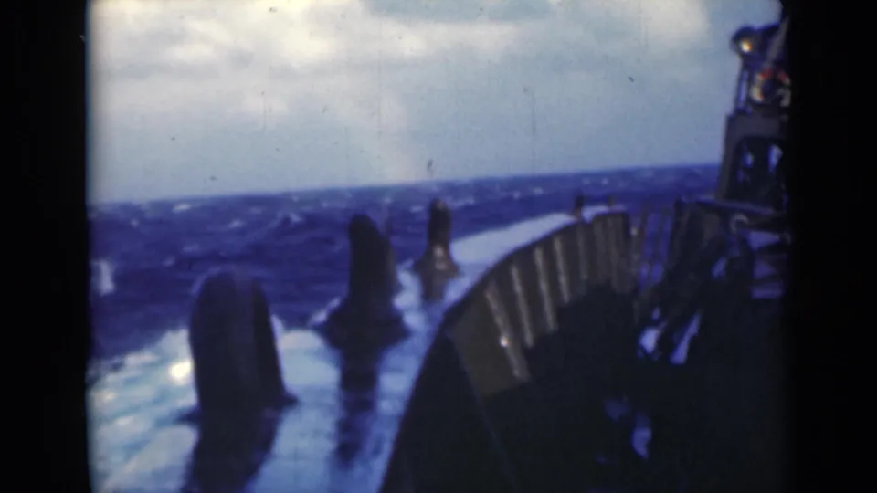 A shipboard 8mm frame shows open blue sea, whitecaps, deck railings, and dark ship structure as the vessel moves through rough conditions.
