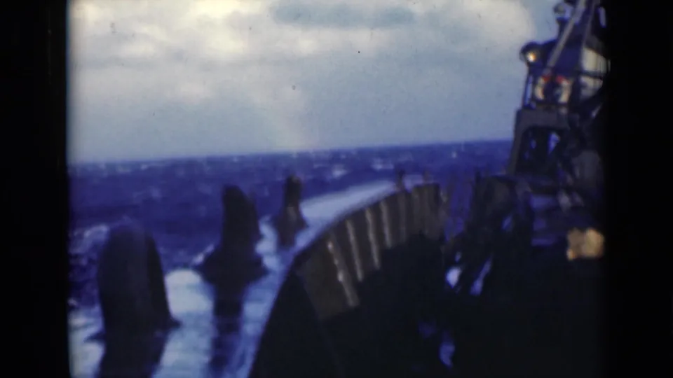 1940s 8mm frame of rough open water viewed from a vessel deck, with railings, deck equipment, and white spray under a cloudy sky.
