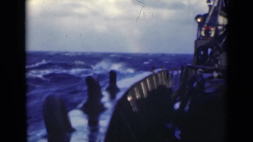 Rough sea surface and whitecaps appear ahead of the vessel, with the deck edge and railings framing the right side of the 8mm frame.