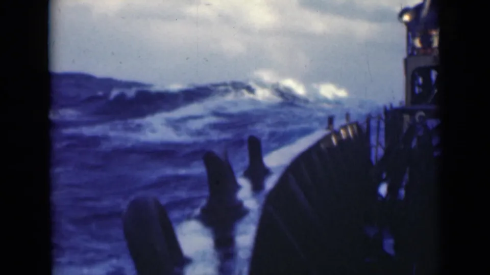A large wave crests beyond the ship deck, with railings and dark deck fittings visible in soft, grainy 8mm color.