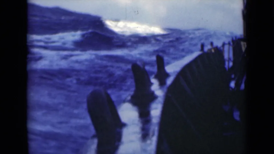 Heavy blue swells and foam fill the frame beside the vessel's rail, with dark deck equipment silhouetted in the foreground.