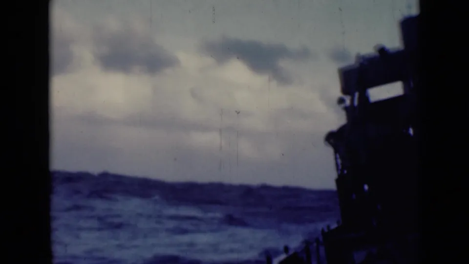 Shipboard 8mm frame showing rolling blue water, low clouds, and a dark vessel silhouette along the right side.