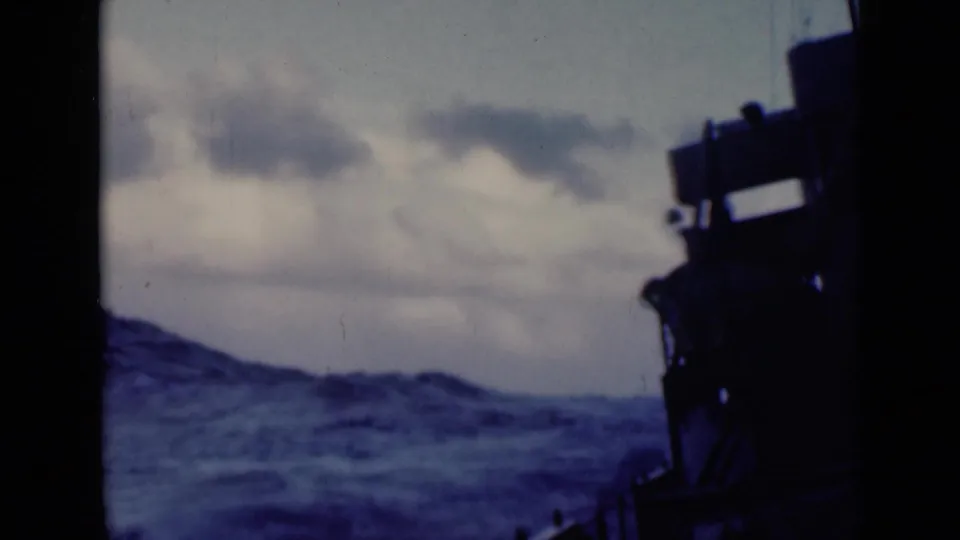 1947 home-movie frame from a moving vessel, with railings, dark superstructure, choppy sea, and overcast sky visible.