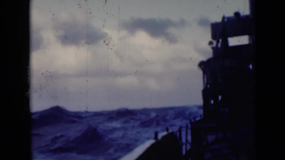Shipboard view of larger rolling waves and whitecaps, with the vessel edge silhouetted against a cloudy horizon.