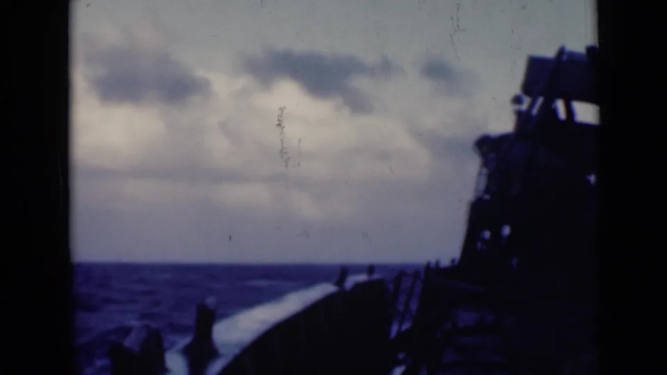 Open sea under a cloudy sky, filmed from a moving vessel with the deck silhouetted along the right edge.
