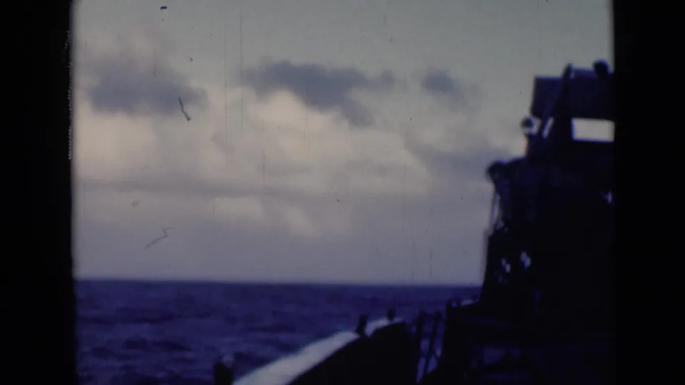 Shadowed ship structure and rail-like forms frame a rough ocean surface beneath low clouds.