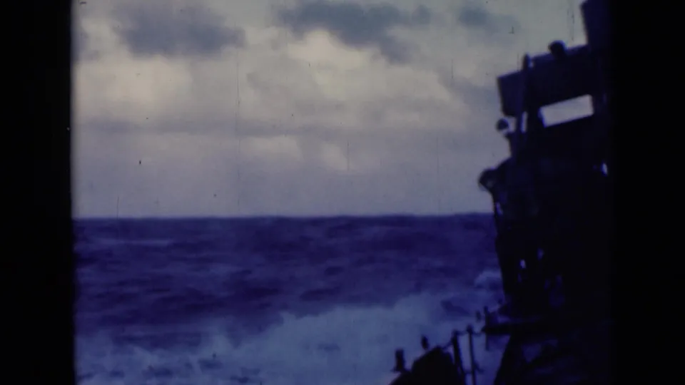 1947 8mm frame of heavy waves under cloudy sky, viewed from a vessel with dark deck structure along the right edge.