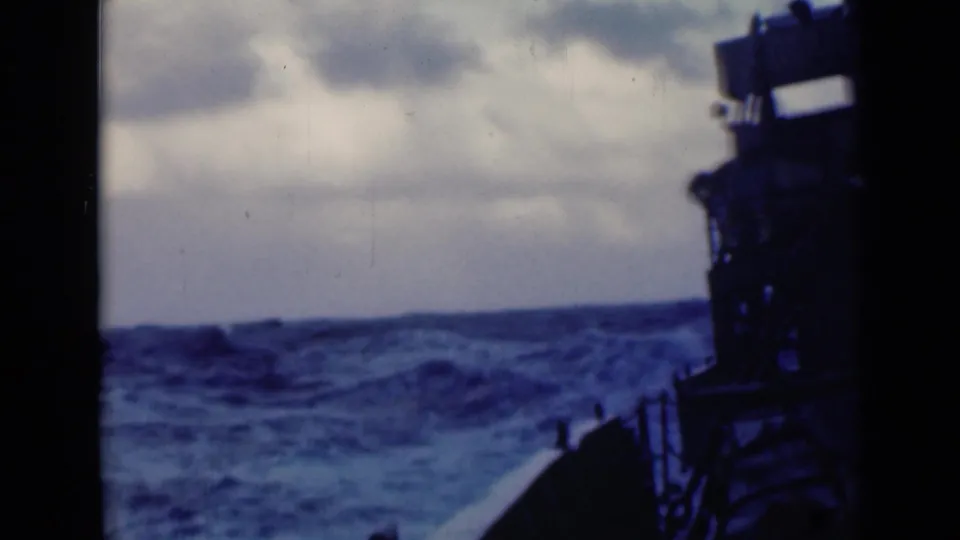 Blue-gray waves and whitecaps stretch across the horizon, with the ship deck and dark vertical structure visible at right.