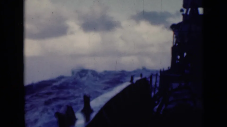 A larger wave breaks near the vessel, with railings and deck structure silhouetted against the stormy sea and cloudy sky.