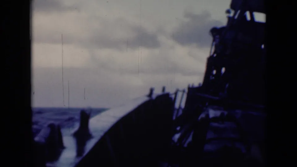 1947 8mm frame showing a deck-level view of a vessel moving through rough sea, with dark superstructure at right and cloudy sky above.