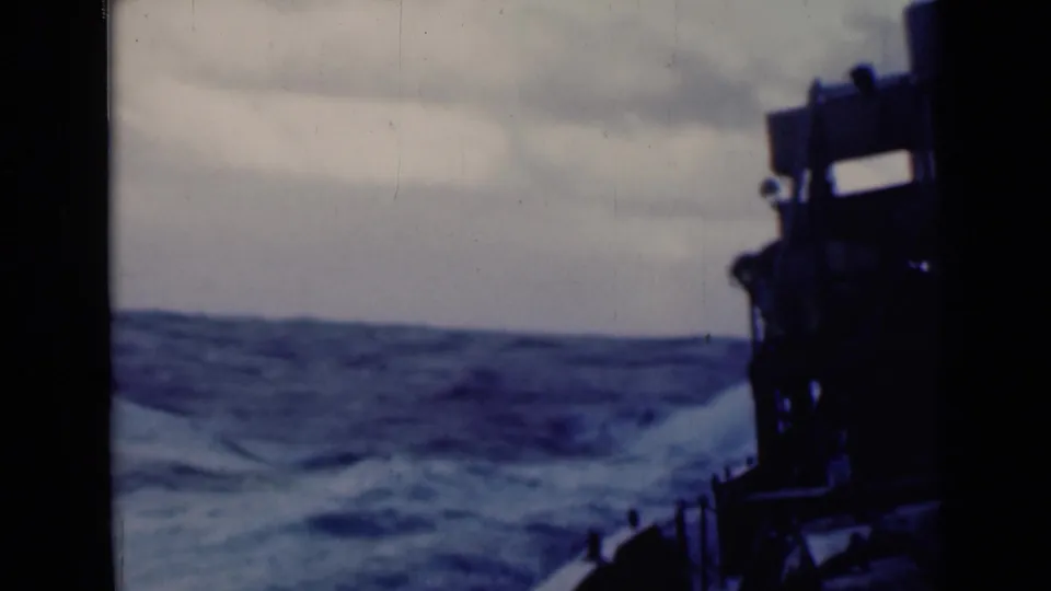 1940s 8mm deck view of rolling sea, with a dark ship structure and railing silhouetted against an overcast sky.