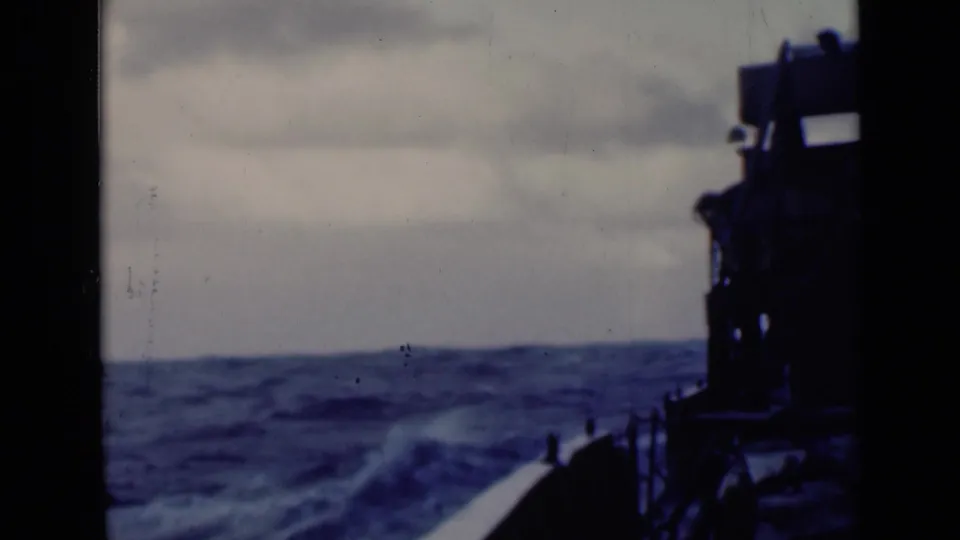 Deck-facing view of choppy sea, with the vessel's forward edge and dark equipment visible against a cloudy horizon.