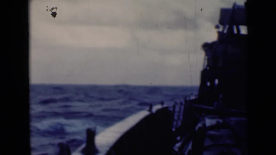 Handheld 8mm frame from a vessel deck, showing open swells, railing posts, and a dark ship structure at right.
