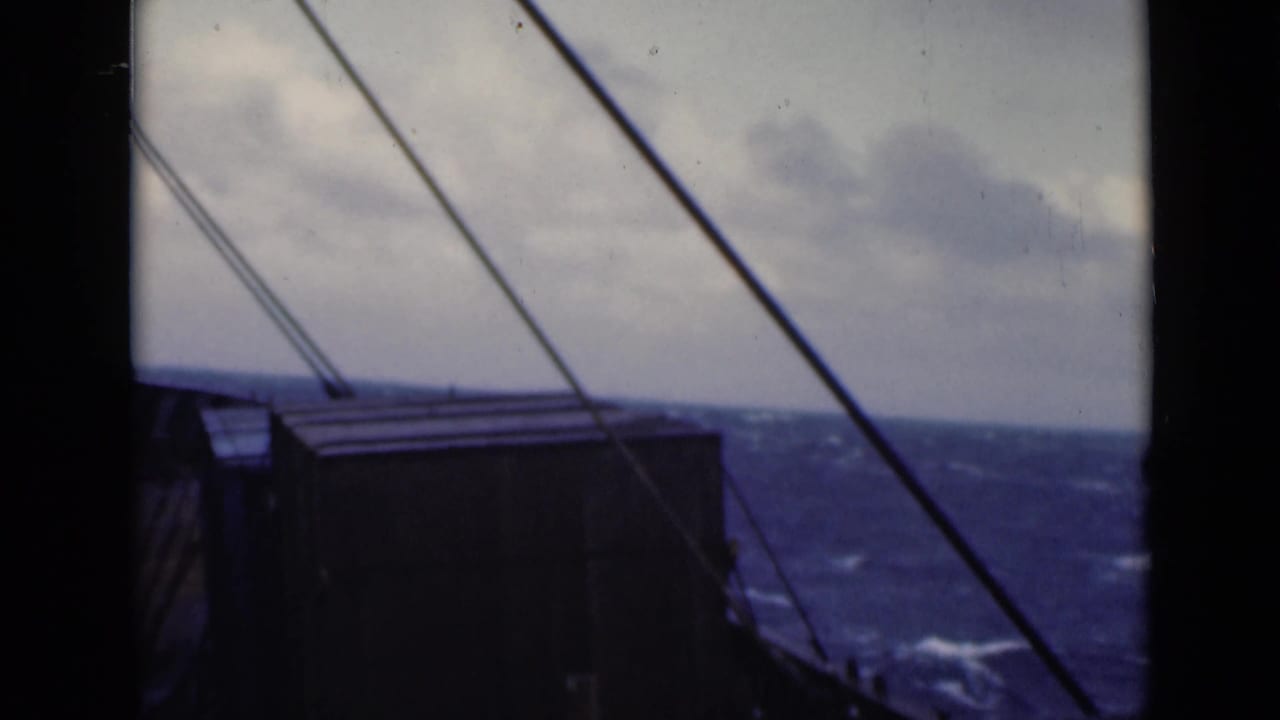 View through a ship window toward rough blue sea, rigging lines, and a dark deck structure.