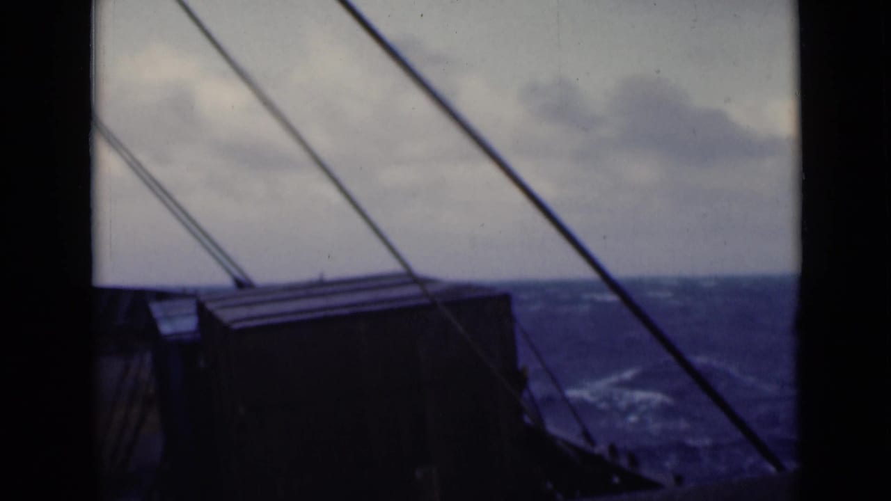 Rough ocean and clouded sky seen past a ship deck structure and diagonal rigging.