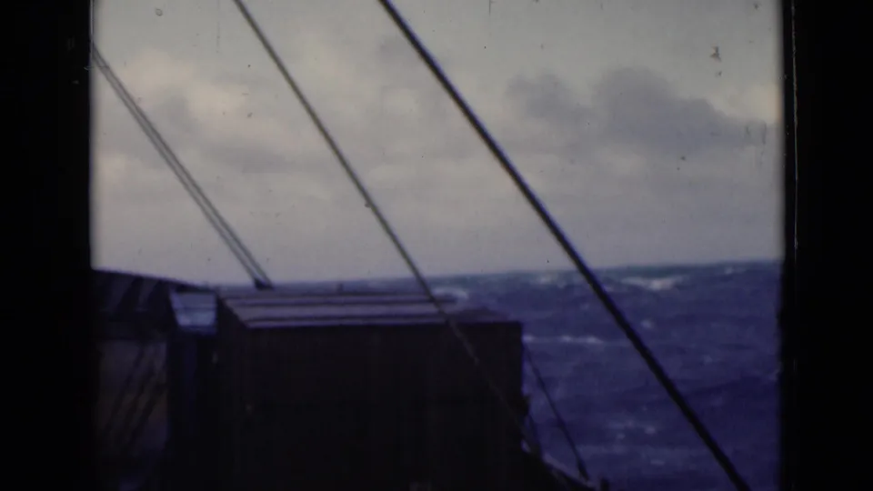 8mm home-movie view from inside or beside a ship window, with deck structure in shadow and rough sea visible beyond.