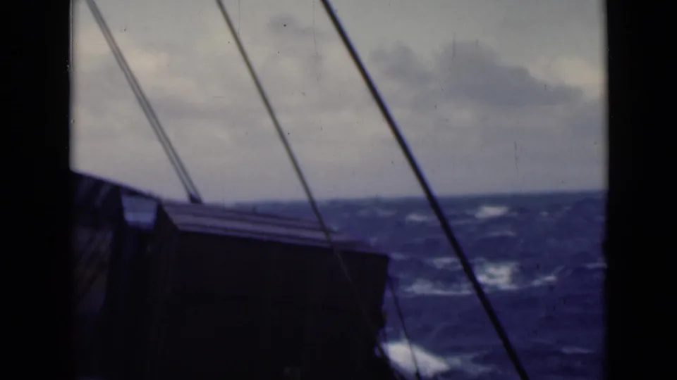 Archival 8mm frame from a cargo ship in rough weather, with rigging crossing the view and whitecaps visible beyond the deck.