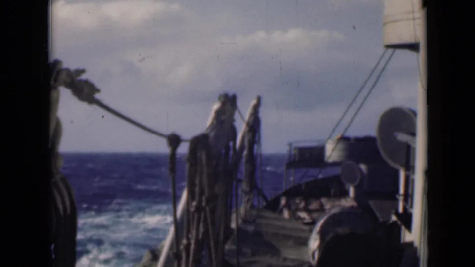 Shipboard 8mm view of rough blue water, foreground rigging, and rounded equipment on deck.