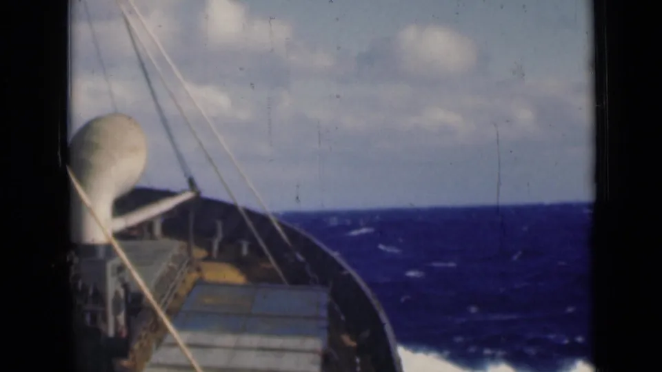 Bow-facing 8mm view of a ship at sea, with rigging lines and a white deck fixture against blue waves and clouds.