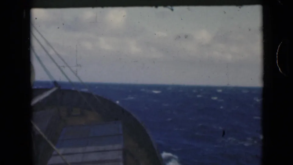 Wide shipboard view from the bow, showing dark blue water, whitecaps, rigging, and a cloudy horizon.