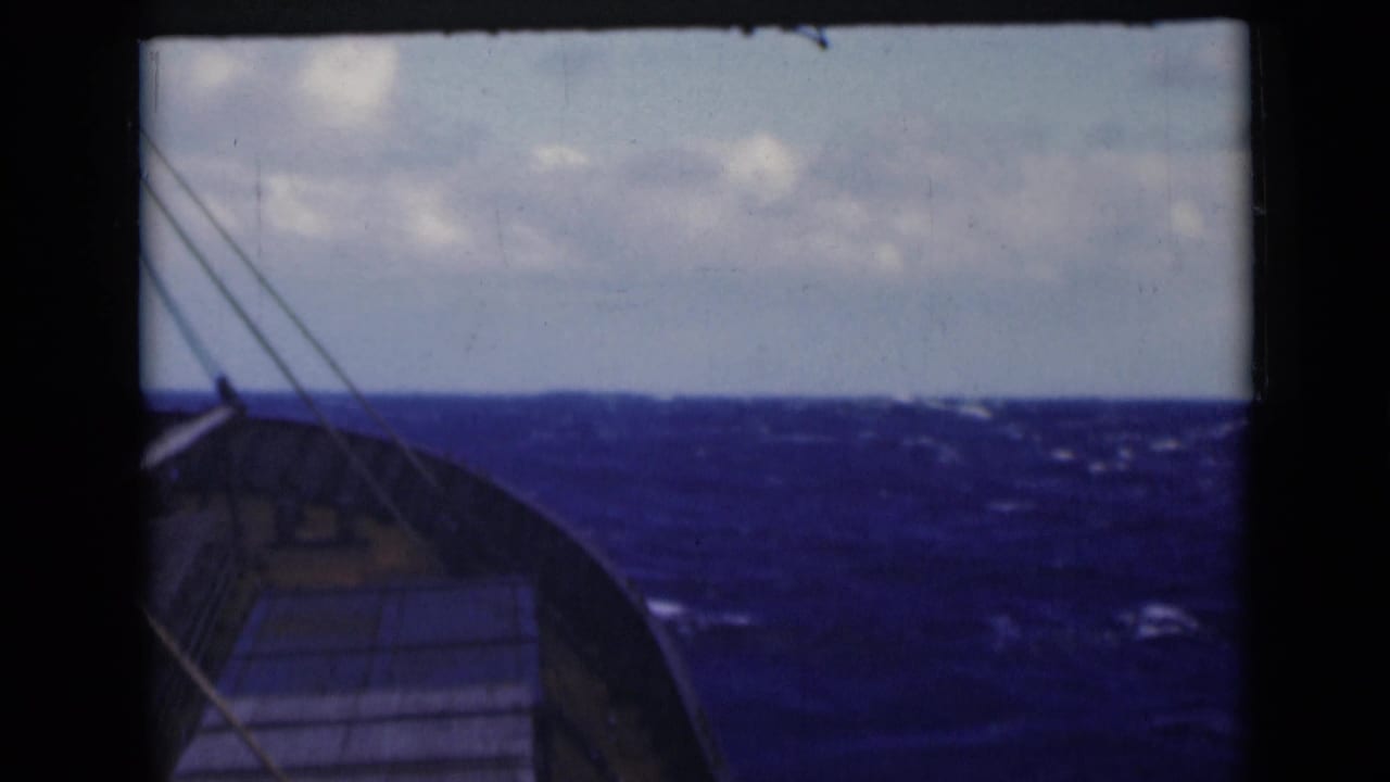 Bow of a vessel points across rolling blue sea with a cloudy horizon.