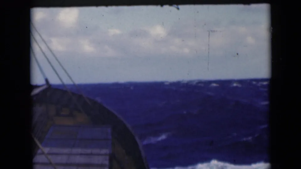 A handheld 8mm shipboard view shows the bow edge beside dark blue waves and a cloudy sky.