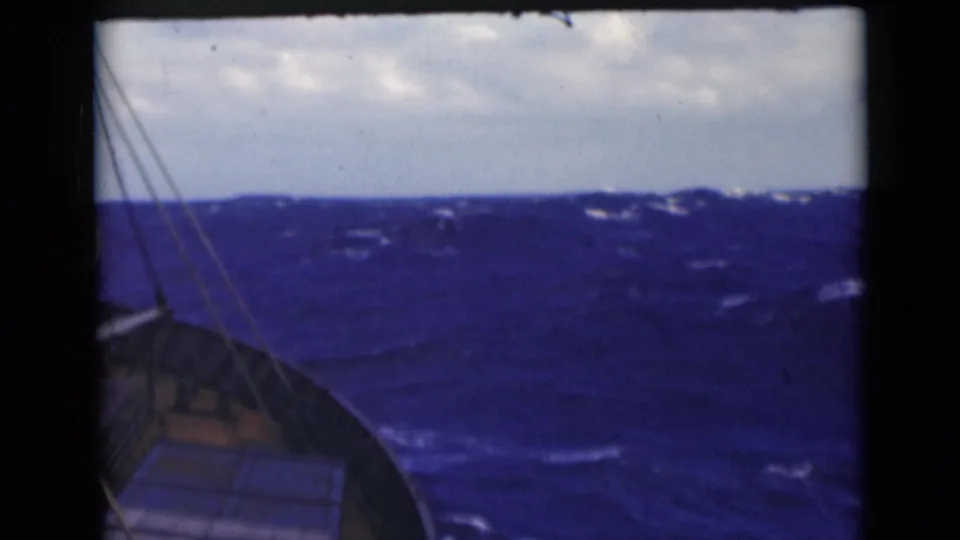 1947 8mm frame showing a deck-side view over choppy blue water with rigging lines, whitecaps, and a cloudy horizon.