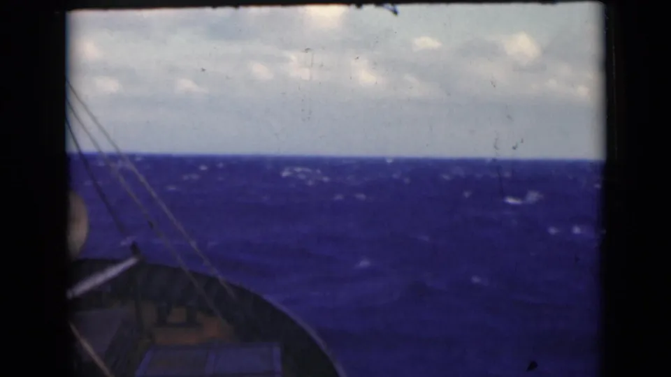 Deck-level 8mm frame showing the boat’s forward edge, rigging lines, rough blue sea, whitecaps, and pale clouds.