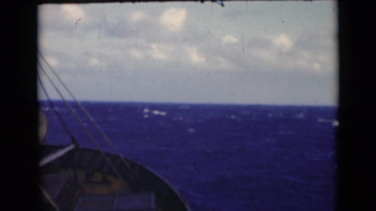 1947 8mm shipboard view over blue water, with bow structure, rigging lines, whitecaps, and cloudy horizon visible.