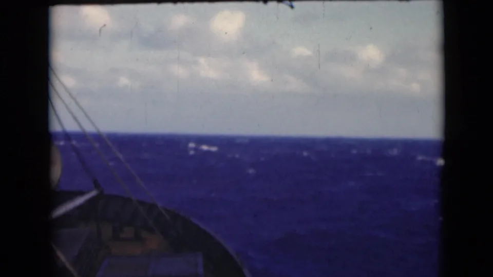 1947 8mm shipboard view over blue water, with bow structure, rigging lines, whitecaps, and cloudy horizon visible.